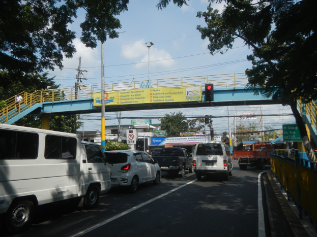 Eine belebte Straße mit Autos und Lastwagen, eine Brücke mit Geländern und Stufen, Laternen, Verkehrszeichen, Schilder mit Text, Bäume, Gebäude und ein bewölkter Himmel.