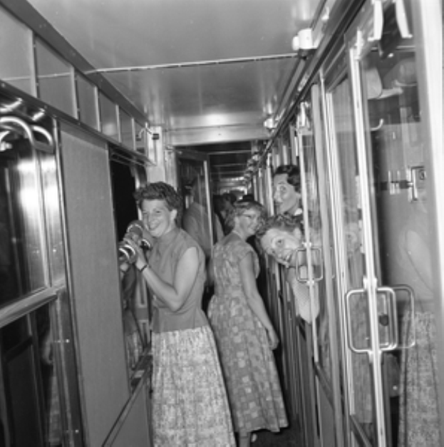 Eine Gruppe von Frauen, die auf einem Zug mit Türen auf beiden Seiten und Lampen oben steht, in Schwarz-Weiß dargestellt.