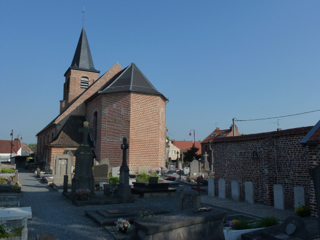 Ein Friedhof mit einer Kirche im Hintergrund, umgeben von Gebäuden, Straßenmasten, Straßenlaternen, Strommasten, Stromkabel, Kraftfahrzeugen auf der Straße, Grabsteinen, Blumensträußen, Bäumen und einem klaren blauen Himmel.