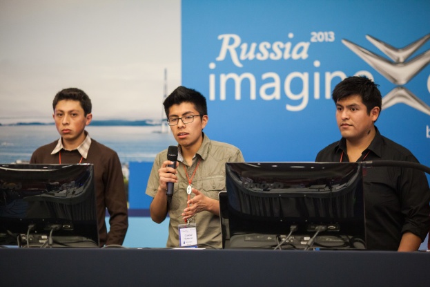 Drei Jungen stehen neben einem Schreibtisch mit zwei Computern, einer spricht in ein Mikrofon, trägt ID-Karten mit roten Markierungen, vor einer Wand mit einer Werbetafel, auf der "Russia Imagine 2013" steht.