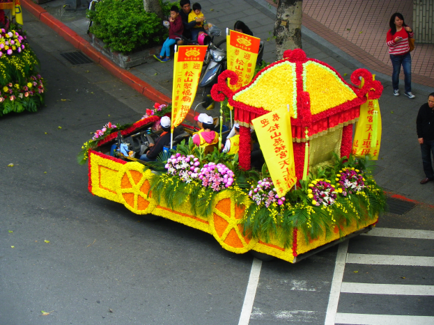 Ein Paradewagen, der mit Blumen und Bannern geschmückt ist und Menschen trägt, bewegt sich auf einem Weg, der von Zuschauern gesäumt ist, mit Bäumen und Pflanzen im Hintergrund.