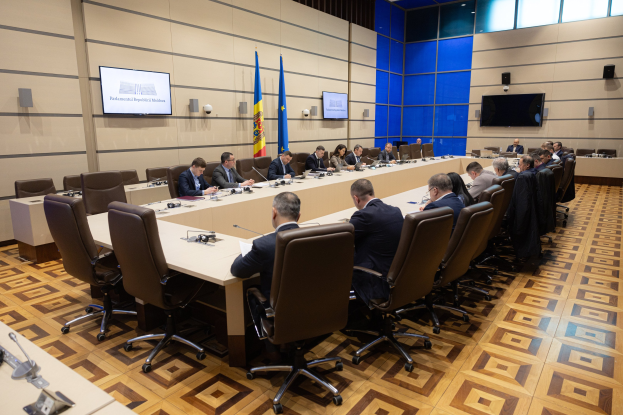 Große Gruppe von Menschen um einen langen Tisch mit Mikrofonen und Papieren in einem Raum mit Flaggen, Fernsehern, Deckenleuchten und Lautsprechern, was auf eine Sitzung des Europäischen Parlaments in Brüssel hinweist.