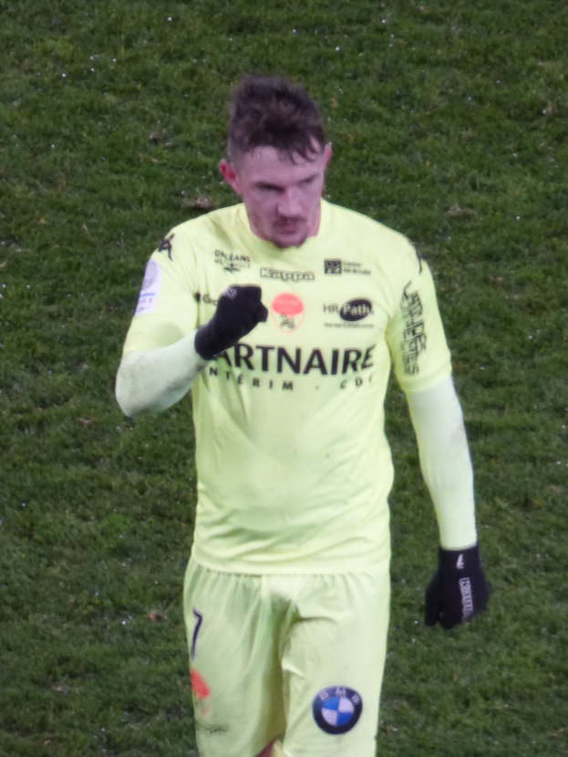 Ein Mann in einem gelben Torwarttrikot, einschließlich eines T-Shirts, Shorts und schwarzer Handschuhe, steht auf einem Fußballfeld.