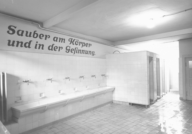 Schwarz-weiß-Foto einer öffentlichen Toilette mit Fliesenboden, Text an einer Wand, mehreren Wasserhähnen, einer Tür und einer Deckenlampe.