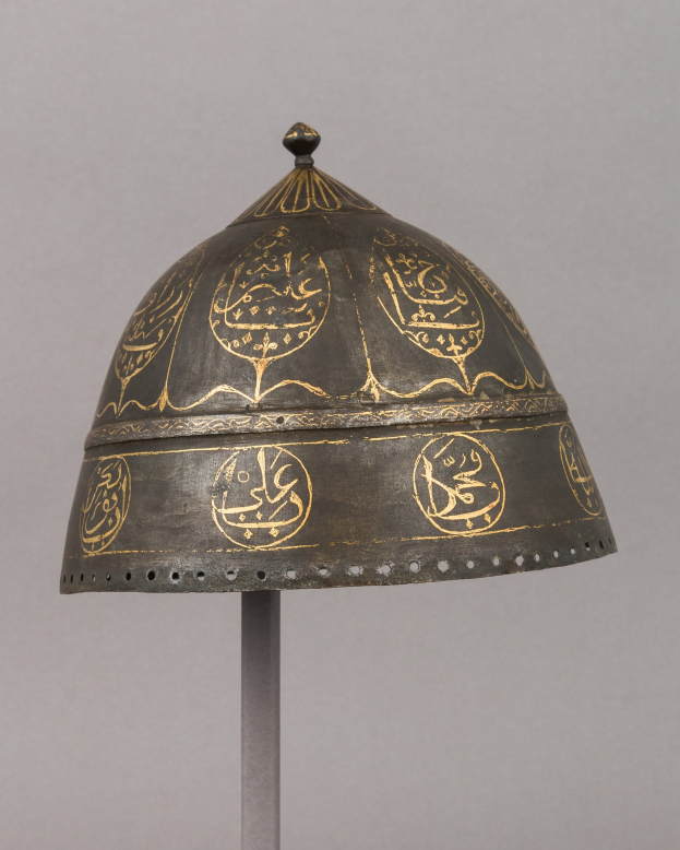 Ein Metallhelm mit detaillierter islamischer Kalligrafie, auf einem Ständer gegen einen weißen Hintergrund gestellt.