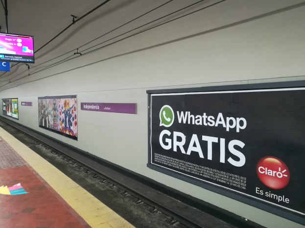 Eine U-Bahn-Station mit einem "WhatsApp Gratis"-Schild an der Wand, ein Bahnsteig mit ein paar Menschen, Schilder mit Text und Bildern und Fernseher, die über dem Bahnsteig angebracht sind.