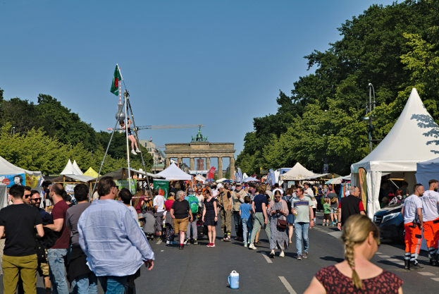 Eine Menge, die eine Straße mit Zelten, Fahrzeugen und Bäumen entlanggeht, mit einem Bogen und einem klaren blauen Himmel im Hintergrund und Flaggenmästen auf der linken Seite.