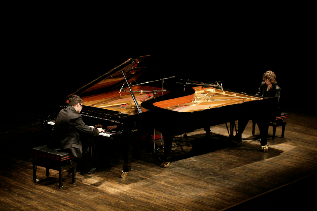 Zwei Personen sitzen an Forte-Pianos auf einer Bühne mit dunklem Hintergrund.