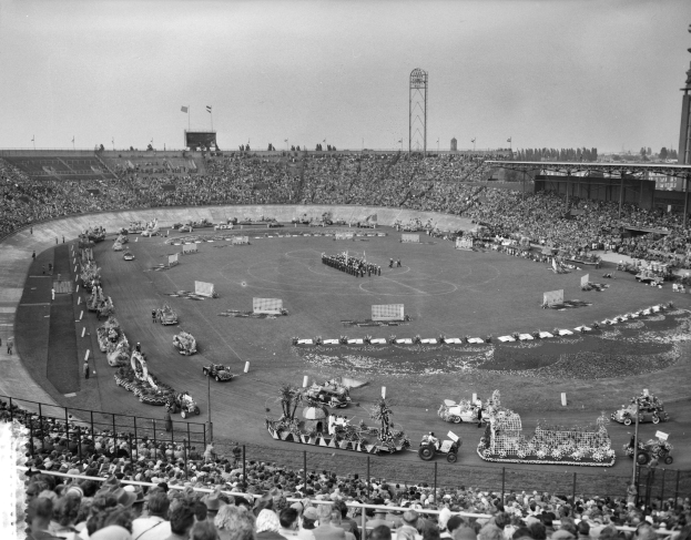 Schwarzes und weißes Bild einer großen Menge, die ein Rennen in einem Stadion mit Fahrzeugen, Zuschauern, Absperrungen, Pfosten, Flaggen, Türmen, Bäumen und Himmel sieht.