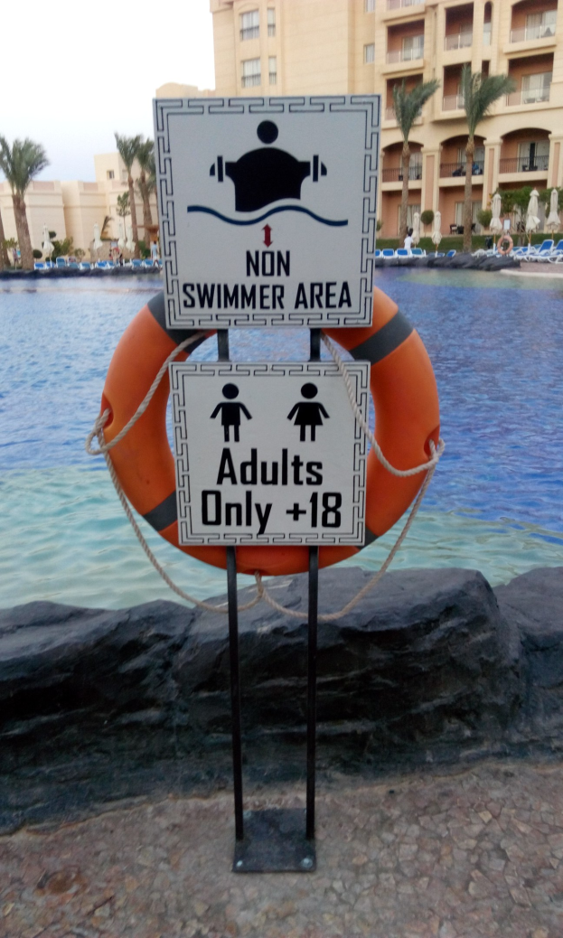 Ein Schwimmbad mit einem "Non Swimmer Area Adults Only"-Schild im Vordergrund, umgeben von Bäumen, Gebäuden und einem klaren blauen Himmel, mit einem bunten Schwimmring in der Mitte des Pools.
