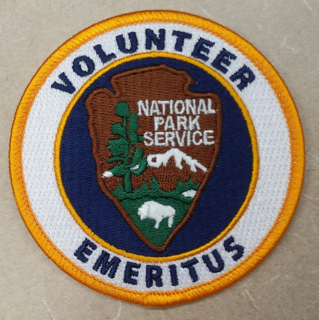 Runder Aufnäher mit "Volunteer National Park Service Emeritus" in fetter schwarzer Schrift auf weißem Hintergrund, umrandet von einem blauen Rand und einem gelben Kreis.