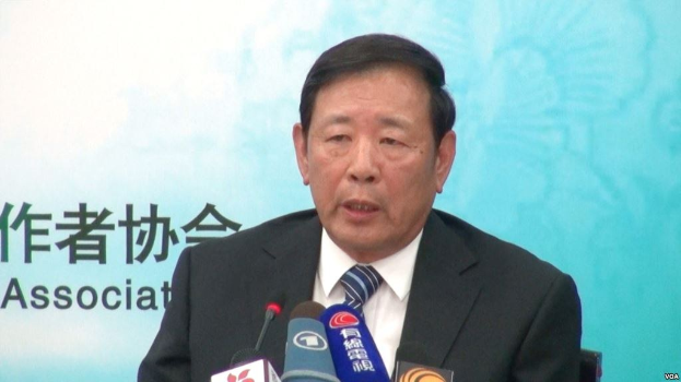 Ein Mann in Anzug und Krawatte sitzt an einem Tisch mit Mikrofonen und spricht während einer Presseveranstaltung mit einem Banner im Hintergrund.