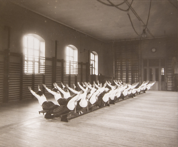 Gruppe von Menschen, die Pilates in einem Fitnessstudio mit einer Fensterwand und einer Uhr im Hintergrund machen.