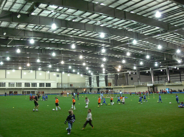 Eine Hallenfußballanlage mit einer Gruppe von Menschen, die mit einem Fußball auf dem Boden spielen, Torpfosten und Beleuchtung auf dem Dach.