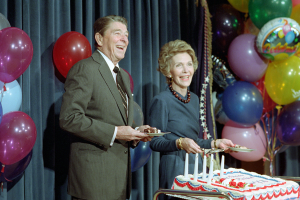 Ein Mann und eine Frau stehen nebeneinander, lächeln und halten Teller in den Händen, vor einem Tisch mit einer Kerzen geschmückten Torte, bunten Ballons und einem Vorhang im Hintergrund, feiern ihren 60. Geburtstag.