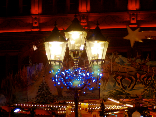 Eine Straßenlaterne in einer Stadt bei Nacht, geschmückt mit Lichtern und Dekorationen, steht vor einem Gebäude und verbreitet eine festliche Stimmung.