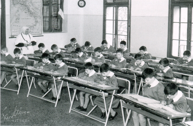 Ein Schwarz-Weiß-Foto eines Klassenzimmers mit Kindern an Tischen, Büchern und Papieren auf den Tischen, einer Uhr und einer Landkarte an der Wand und Fenstern im Hintergrund.