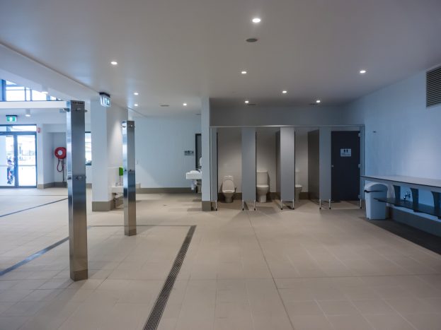 Großes öffentliches WC mit mehreren Toiletten, Säulen, einem Müllbehälter, einer Tür, einem Schild, einem Feuerlöscher und Deckenleuchten.