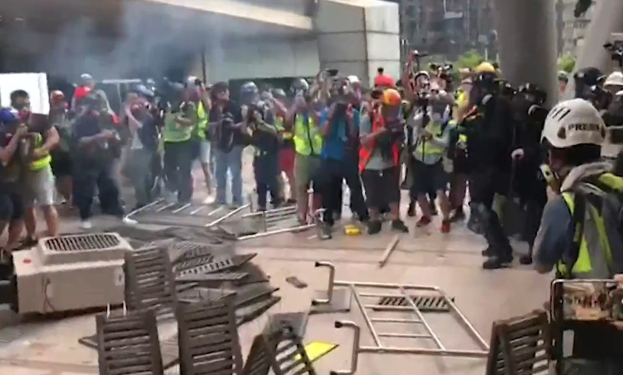 Menschen in Helmen und Jacken stehen nahe einem rauchenden Gebäude, einige halten Kameras, mit verstreuten Metallobjekten und Stadtgebäuden im Hintergrund.