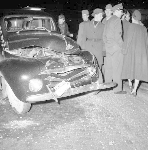 Schwarzes und weißes Bild eines schwer beschädigten Autos am Straßenrand mit eingedellter Motorhaube und zersplitterten Fenstern, umgeben von Menschen, die die Szene begutachten.