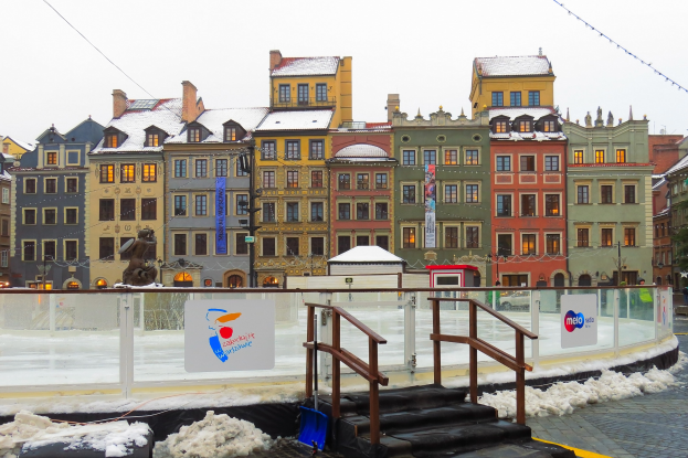Eislaufbahn in einem Stadtplatz umgeben von Gebäuden, Treppen, Geländern, Informationsschildern, schneebedeckter Erde, Laternen, Bäumen und einem Himmel als Hintergrund.