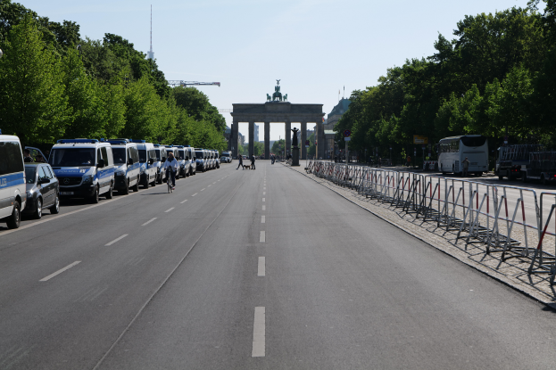 Eine Reihe von Polizeiwagen auf einer Straße vor dem Brandenburger Tor in Berlin, Deutschland, mit Menschen auf Fahrrädern und Stehenden, Barrieren, Bäumen und einem Bogen mit Statuen im Hintergrund.