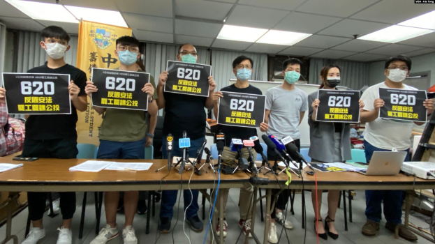 Eine Gruppe maskierter Personen mit Schildern versammelt sich um einen Tisch mit Mikrofonen, Papieren und Laptops, um gegen das Auslieferungsgesetz in Hong Kong zu protestieren.