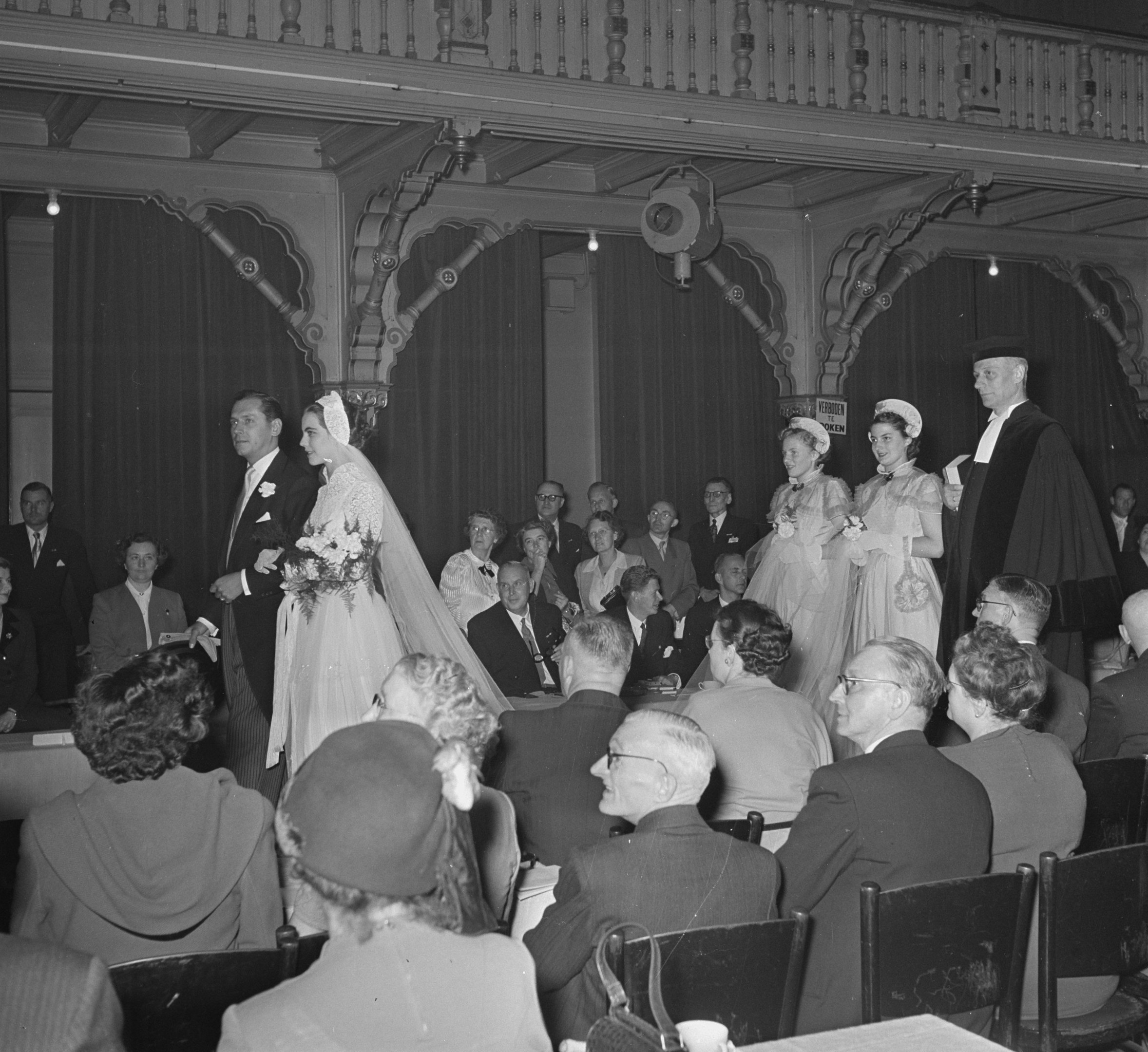 Schwarz-weißes Foto einer Hochzeitszeremonie mit Gästen auf Stühlen und Stehplätzen, Blumensträuße, Tischen mit Tassen und Vorhängen mit Deckenleuchten im Hintergrund.