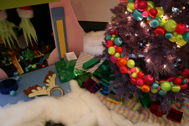 Drinnen steht rechts ein geschm├╝ckter Weihnachtsbaum mit Lichterkette und Schmuck, in der Mitte Geschenkboxen und Deko, oben links eine H├╝tte mit Deko und unten Wolle und Watte.