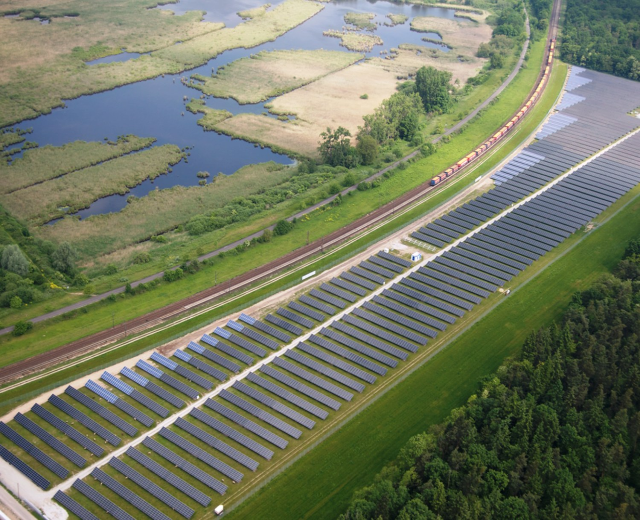 Luftaufnahme eines Solarparks mit Solarpanelen in einem Feld, umgeben von Bäumen, Gras, Pflanzen und Wasser, mit einem Zug auf einer nahen Bahnstrecke.