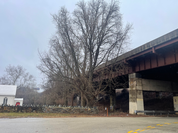 Eine Brücke über eine Straße in Reparatur mit einem Baum in der Mitte, umgeben von verstreuten Werkzeugen, Materialien, Steinen, Gras, einem Haus, einem Schild, einem Straßenpfahl, einer Gruppe von Bäumen und einem bewölkten Himmel.