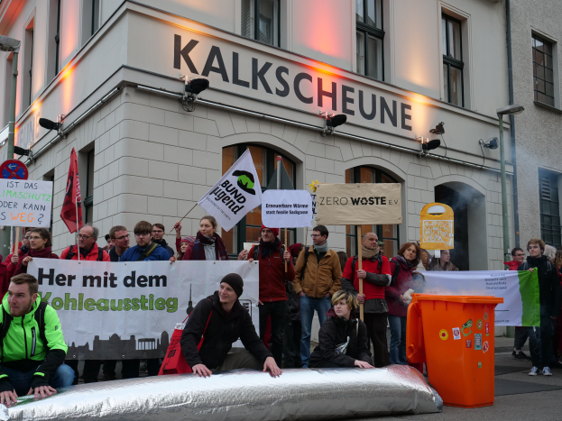Eine Gruppe von Menschen mit Schildern und Plakaten steht vor einem Gebäude, zwei Personen sitzen im Vordergrund und ein Müllcontainer rechts, während einer Demonstration in Deutschland, mit Gebäuden, Fenstern, Lichtern und Schildern im Hintergrund.