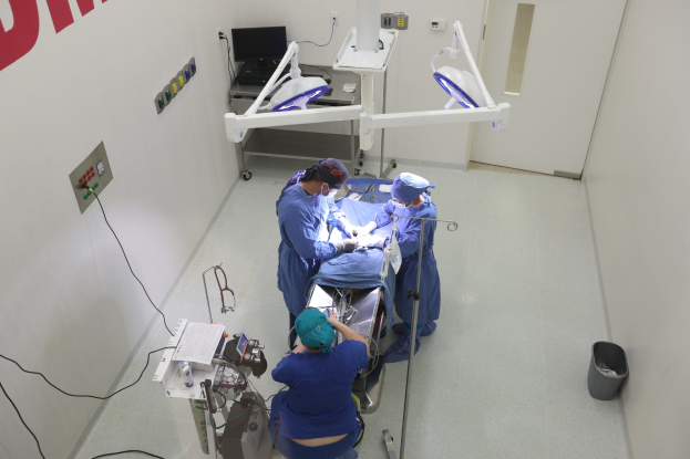 Chirurgen führen eine Operation an einem Patienten in einem Operationssaal mit medizinischer Ausrüstung, einem Monitor und einer Wand mit Text auf der linken und einem Objekt auf dem Boden auf der rechten Seite durch.