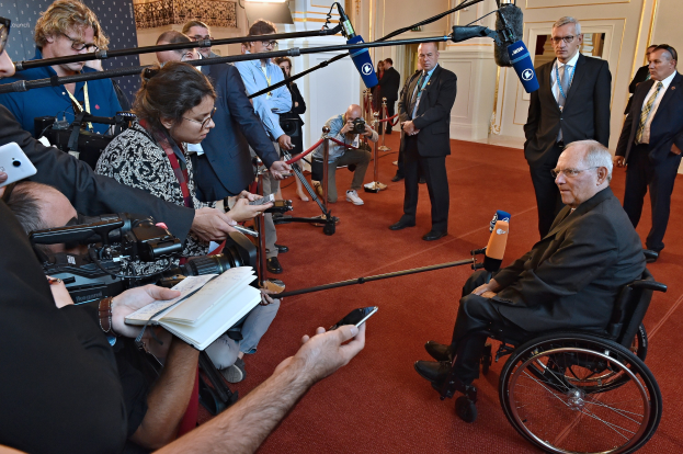 Ein Mann im Rollstuhl spricht mit Reportern, die Kameras, Mikrofone und Handys halten, vor einem Banner, Lichtern und einer Wand.