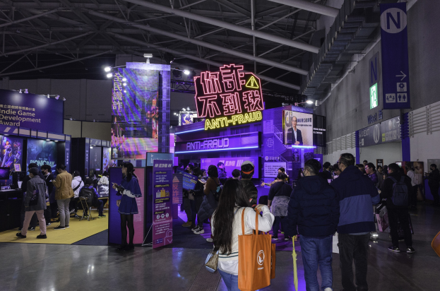 Eine große Gruppe von Menschen steht vor einem Messestand, der als "Indie Game Development Award 2019" gekennzeichnet ist, wobei einige Taschen und Platten, Bildschirme und Lichter im Hintergrund zu sehen sind.