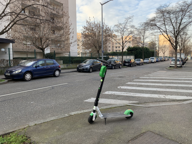 Elektrisches Scooter auf der Straßenseite mit Fahrzeugen, Laternen, Bäumen, Gebäuden und einem bewölkten Himmel im Hintergrund.
