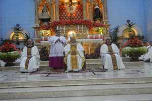 Eine Gruppe von Priestern sitzt auf den Stufen einer Kirche, umgeben von einem Teppichboden, Blumensträußen, Statuen, Kerzen mit Ständern und anderen Gegenständen, wobei die Kirchenwände im Hintergrund sichtbar sind.