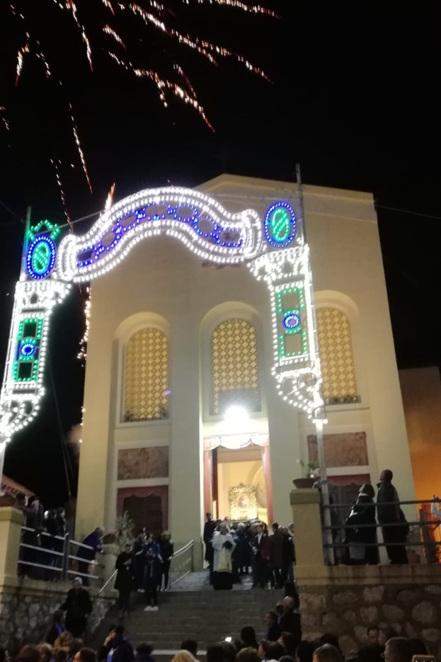 Menschen vor einem Gebäude mit einem bogenförmigen, lichtgeschmückten Eingang, Treppe, Geländern und Topfpflanzen, die Silvester mit Feuerwerk im Himmel feiern