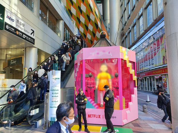 Eine Gruppe von Menschen steht vor einem modernen Gebäude mit Glaswänden, Säulen und Texttafeln, einige tragen Masken, mit einer Rolltreppe im Blick; ein pinker Kasten in der Mitte zeigt Schaufensterpuppen und trägt die Aufschrift "Netflix's new Netflix booth in Tokyo."