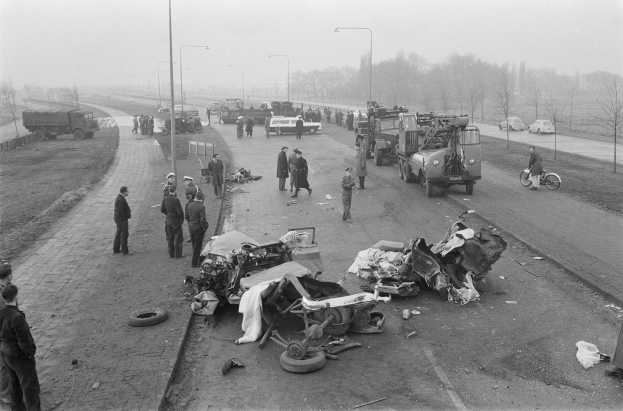 Schwarz-weiß-Szene eines Autounfalls am Straßenrand mit mehreren Fahrzeugen und einer Gruppe von Menschen drumherum, Laternenpfähle, Bäume und Himmel im Hintergrund.