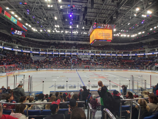 Eine große Menschenmenge in einer Hockeyarena, mit einigen Sitzenden und anderen Stehenden, die ein Spiel unter hellen Lichtern und Bannern beobachten.