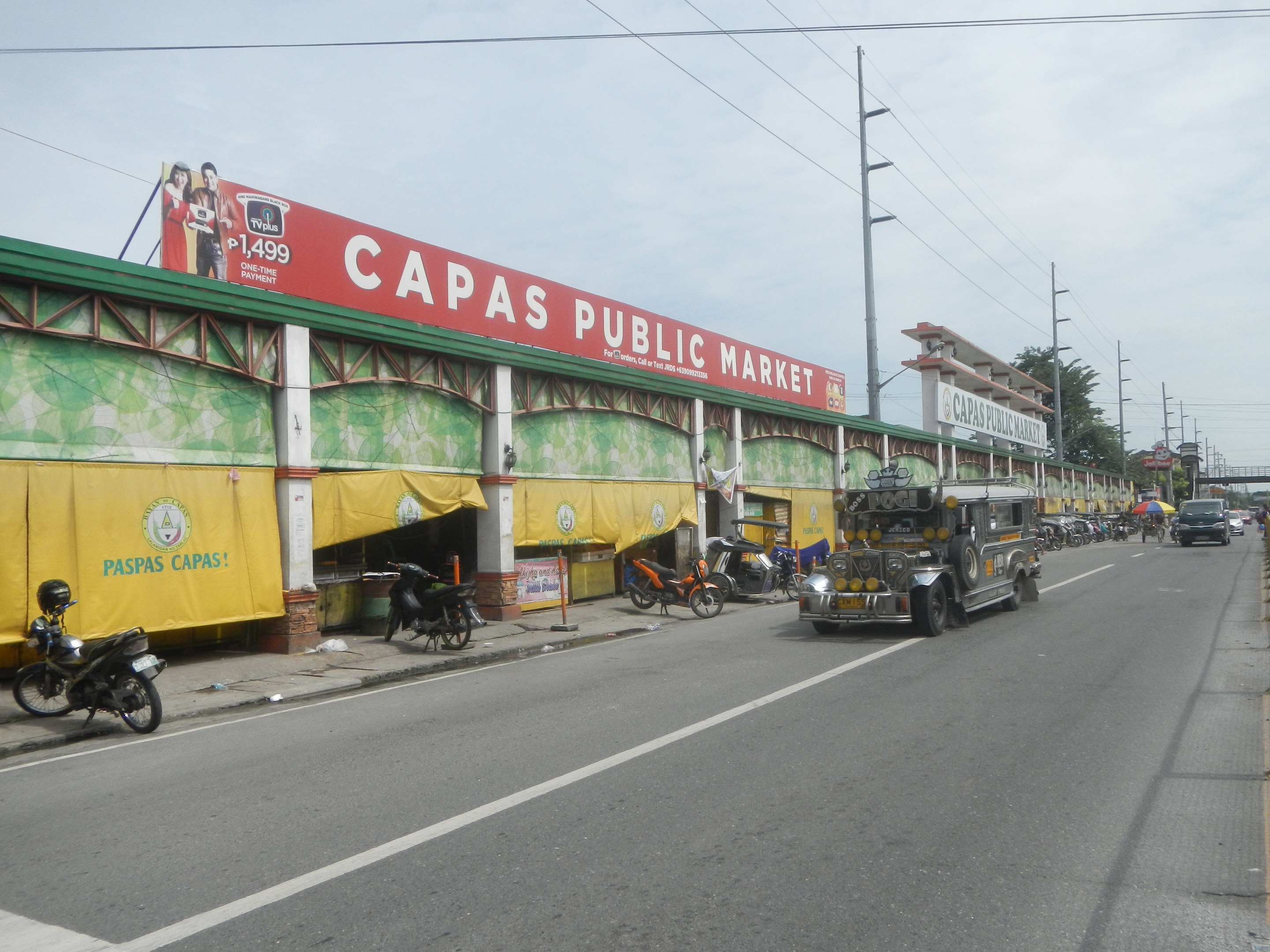 Eine belebte Straße mit Fahrzeugen, einem Gehweg, Strommästen, Gebäuden, Bäumen und einem bewölkten Himmel, mit einem Gebäude mit der Aufschrift "Capas Public Market" im Vordergrund.