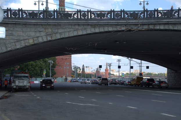 Eine Brücke überspannt eine vielbefahrene Straße mit darunterfahrenden Fahrzeugen; Fußgänger stehen auf dem Gehweg, und im Hintergrund sind Laternenpfähle, Ampeln, Bäume, Gebäude und der Himmel zu sehen.