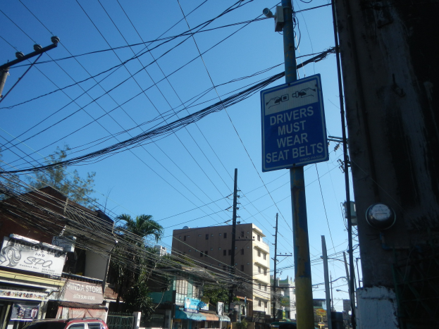 Stadtstraße mit fahrenden Autos, Strommäste mit Drähten, Gebäude, Bäume und Namenschilder; ein Pfahlschild zeigt "Fahrer müssen Sitzgürtel tragen" gegen einen sichtbaren Himmel.