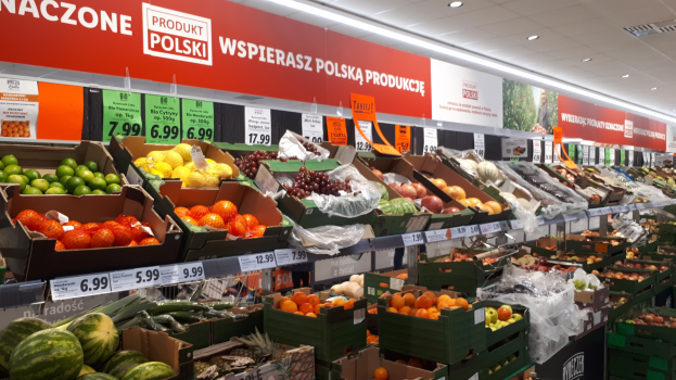 Ein Geschäft mit Kartonagen voller Obst und Gemüse, jeweils mit Preisschildern, an der Wand montierten Tafeln mit Text und Bildern und Deckenleuchten.