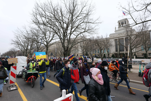 Eine große Gruppe von Menschen nimmt an einer Protestdemo auf einer Straße in Washington, D.C. teil, einige halten Schilder und Banner, andere fahren Fahrräder, und Schilder, Bäume und ein klarer blauer Himmel im Hintergrund.