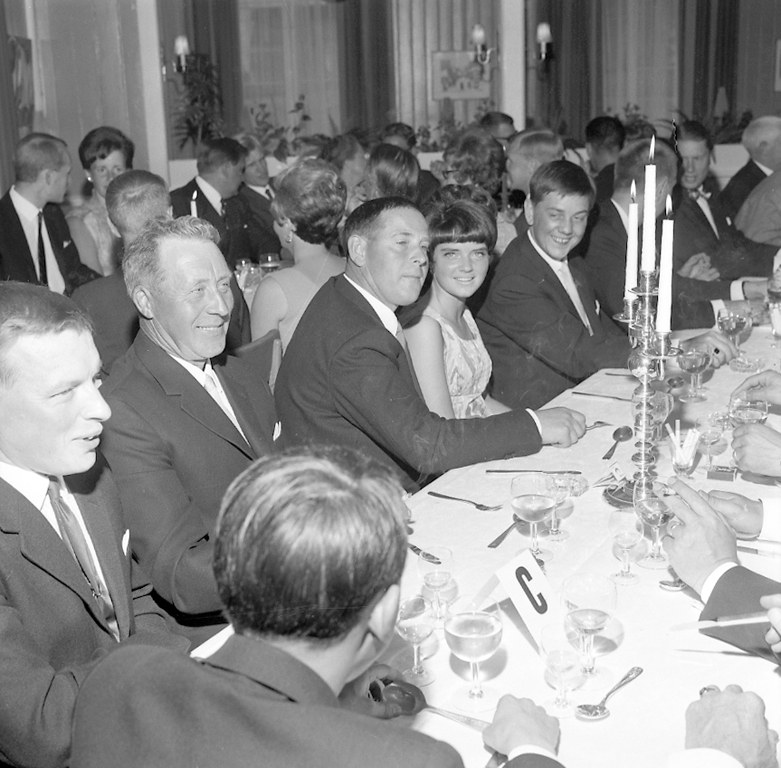 Eine Gruppe von Menschen sitzt um einen langen Tisch mit Gläsern, Besteck und Kerzen, die in einem warm beleuchteten Raum mit Vorhängen im Hintergrund strahlen.