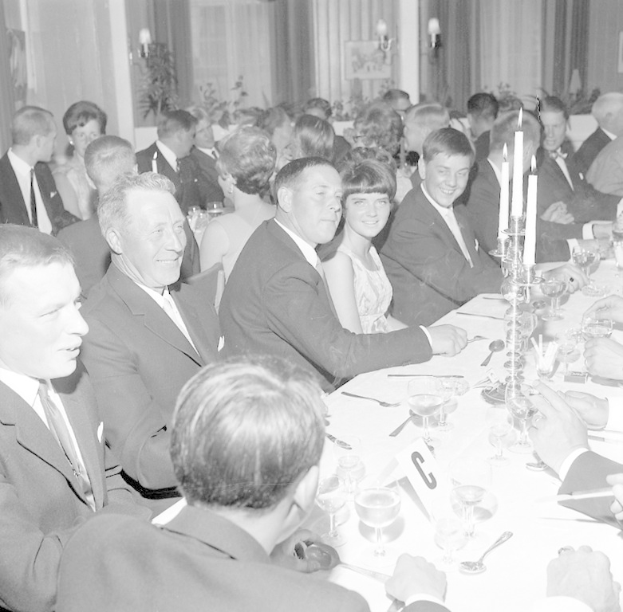 Eine Gruppe von Menschen sitzt um einen langen Tisch mit Gläsern, Besteck und Kerzen, die in einem warm beleuchteten Raum mit Vorhängen im Hintergrund strahlen.