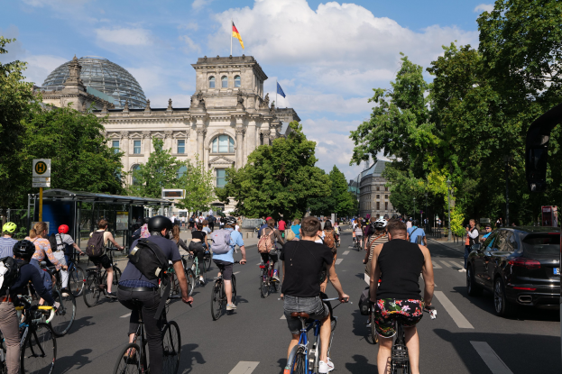 Gruppe von Menschen, die auf Fahrrädern eine von Bäumen gesäumte Straße in Berlin entlangfahren, mit Gebäuden, einem Bushaltestellen auf der rechten Seite und einer Fahne, die auf einem der Gebäude weht, unter einem bewölkten Himmel.