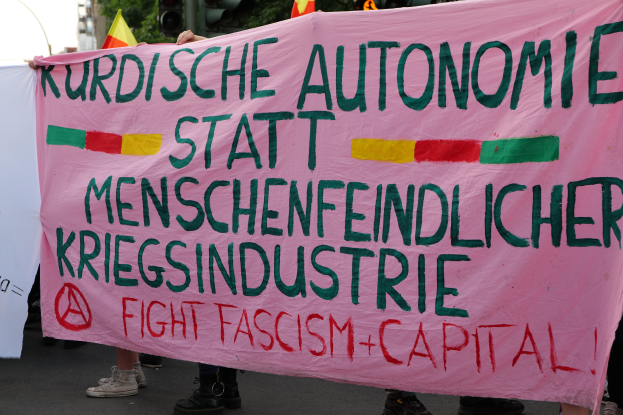 Eine Gruppe von Menschen steht auf der Straße und hält ein pinkes Banner mit der Aufschrift "Kurdische Autonomie Statt Menschenfeindlicher Kriegsindustrie Fight Fascism Capital", mit Verkehrsampeln, Flaggen, Bäumen, Gebäuden und einem klaren blauen Himmel im Hintergrund.